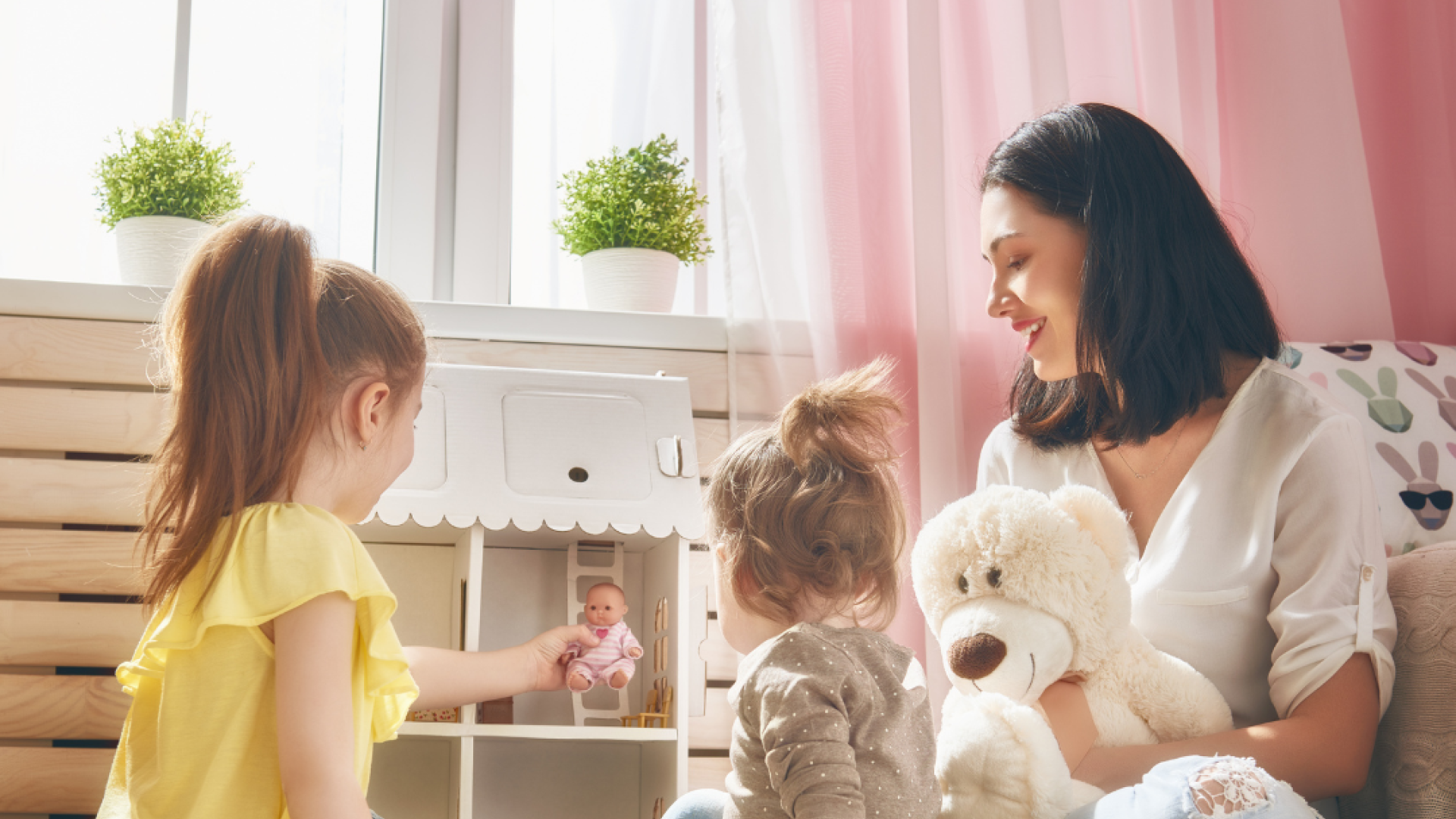 A woman and her two children play with a dollhouse and a teddy bear
