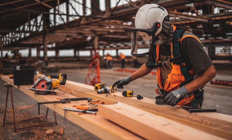 Construction worker with a hard hat and face shield on, measuring wood planks.