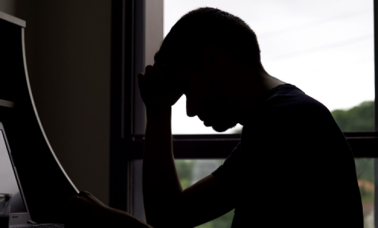 A man in shadow sits at a desk rubbing his head