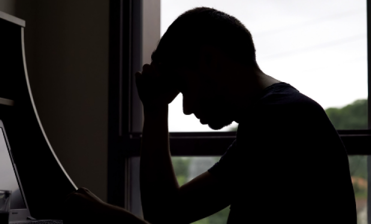 A man in shadow sits at a desk rubbing his head 