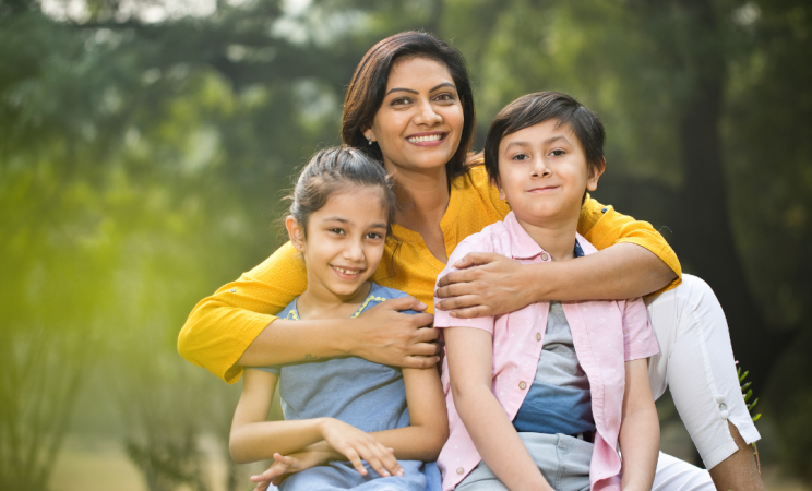 A mother hugs her two young children as they sit outside in nature