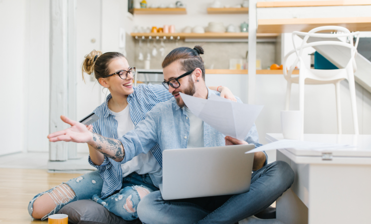 A man and a woman sit on the kitchen floor with a laptop and papers discussing their taxes