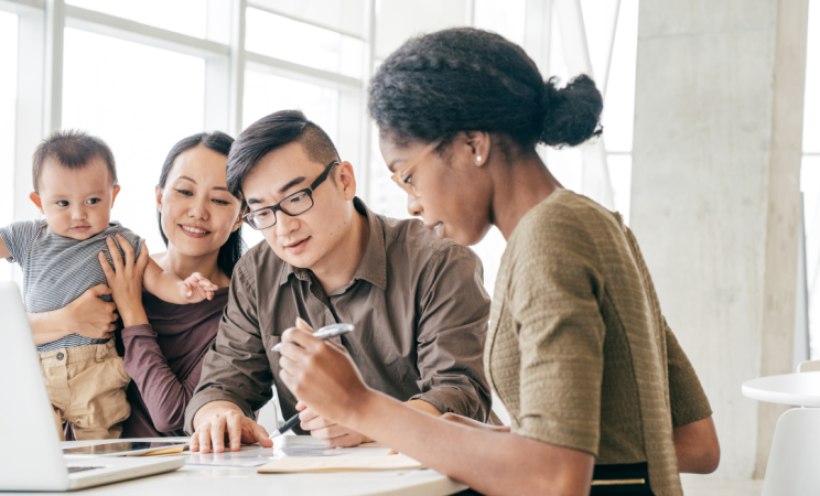 A woman holding her baby sits next to a man and woman as they go over a tax return on the kitchen table.