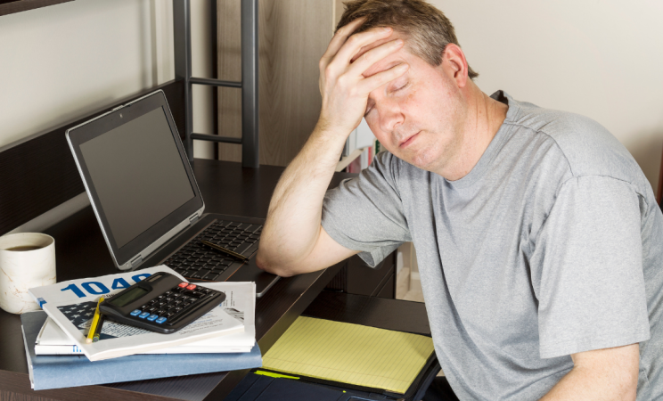 A man holds his hand in his head in frustration with a calculator and tax form on a wooden desk he is sitting at. 