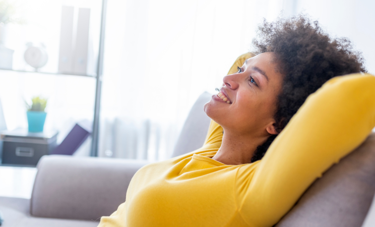 A woman in a yellow sweater leans back on a grey couch with her hands behind her head with a relaxed and happy expression on her face