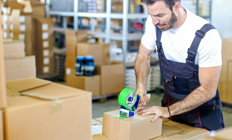 A man in a workshop apron closes a cardboard package with a roll of package tape.