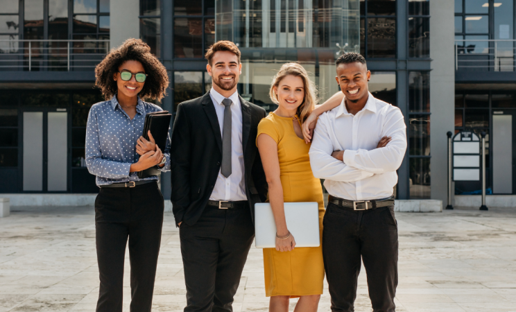 Four young people in professional wear pose for a picture in front of a large glass building