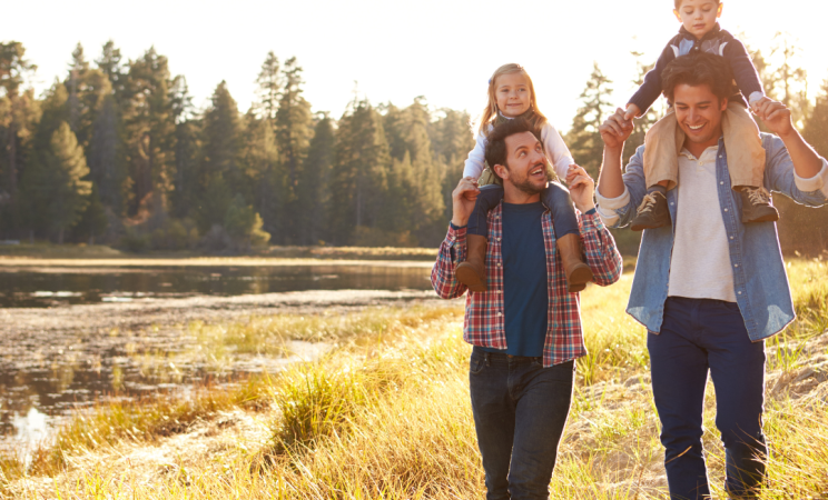 Two men hike through a wood lined field. Each man has a small child on his shoulders. 