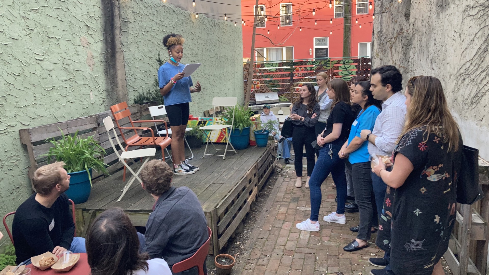 People gather in a garden to watch a young woman speak on a wooden stage. 