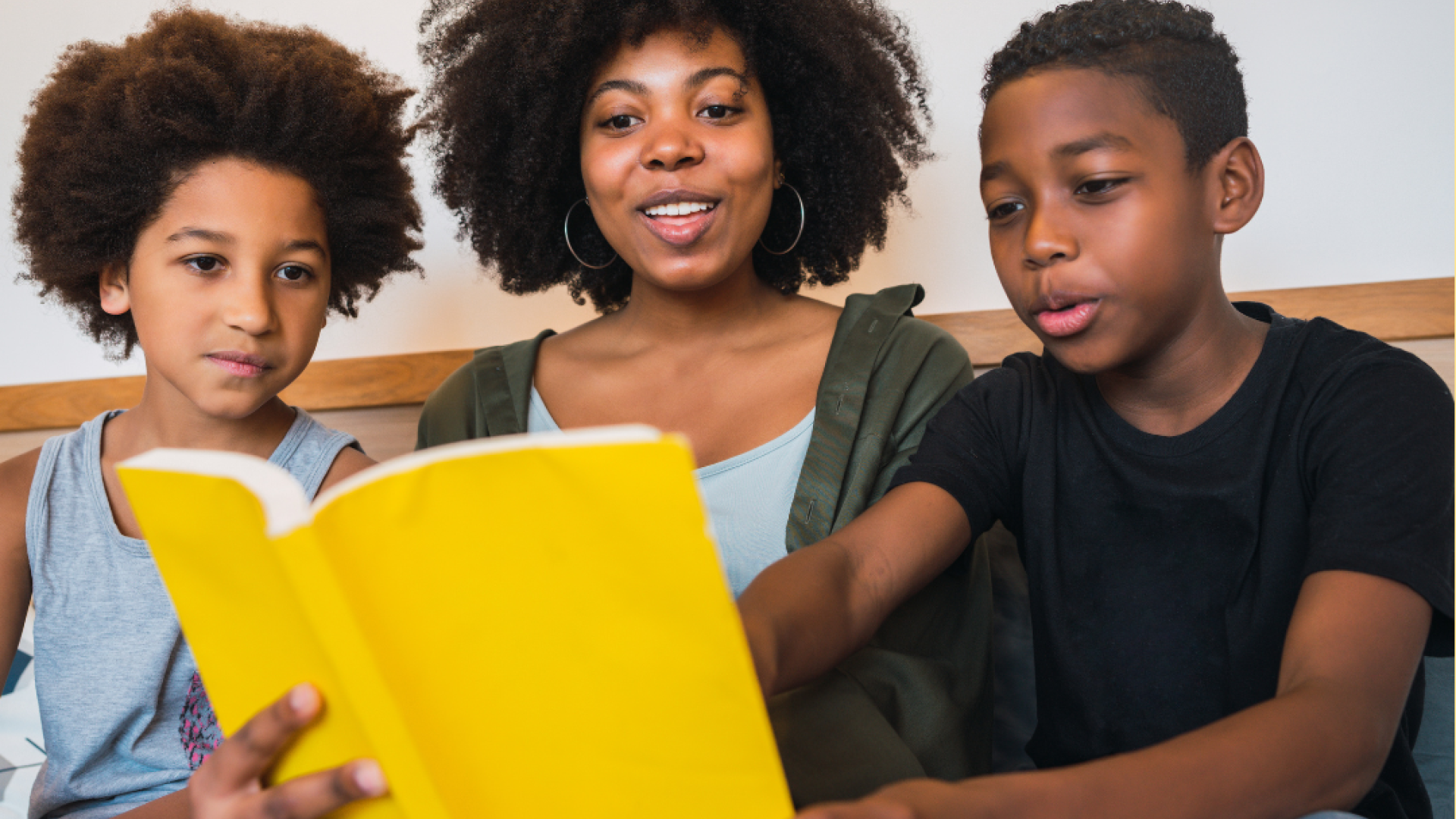 A mother reads a yellow book to her two elementary school aged children. 