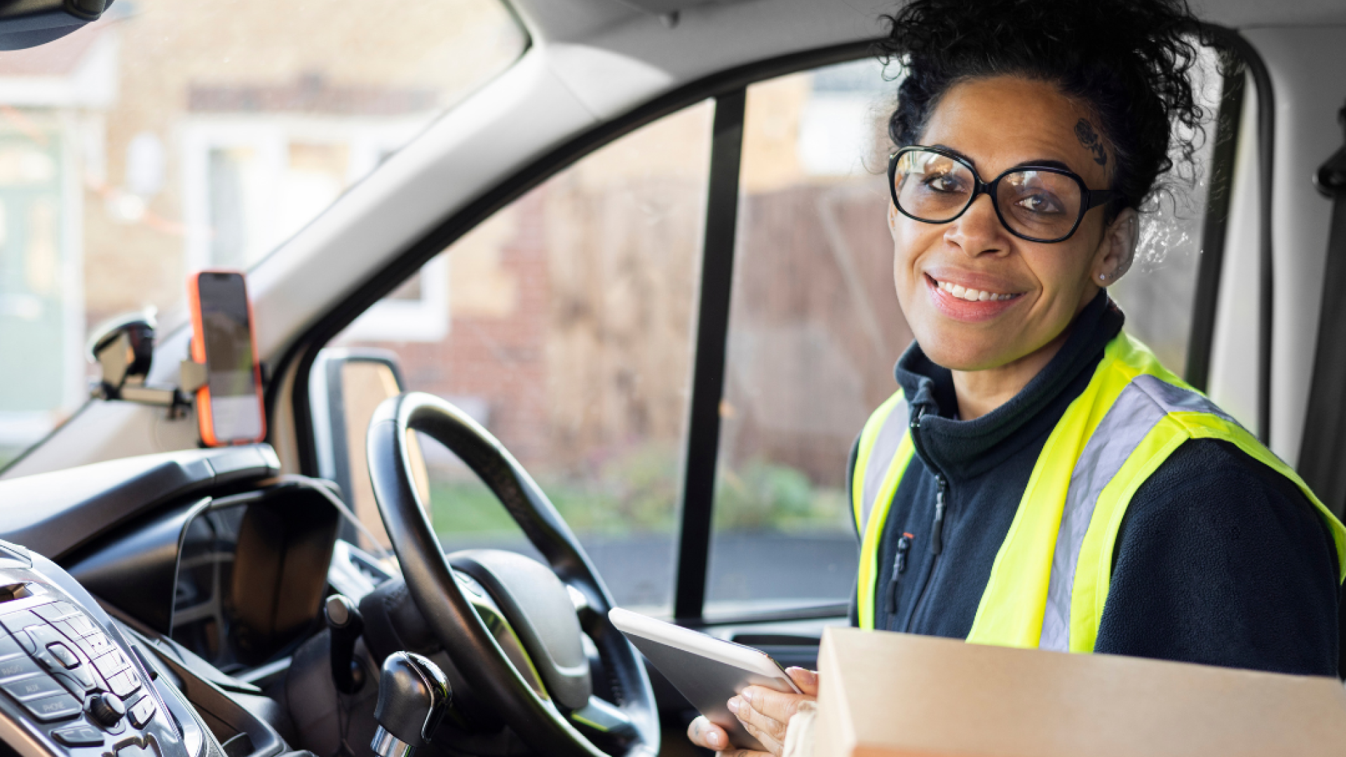 A woman in a yellow safety vest sits in the cab of a delivery truck smiling at the camera