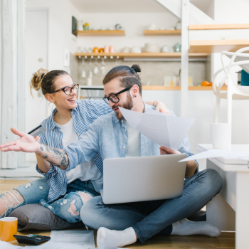 A man and a woman sit on the kitchen floor with a laptop and papers discussing their taxes