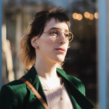 A woman in a green coat and wire framed glasses looks to her left meaningfully