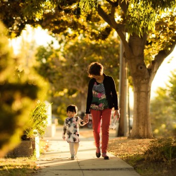 A woman holds her toddlers hand as they walk down a tree lined sidewalk