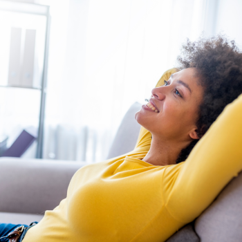 A woman in a yellow sweater leans back on a grey couch with her hands behind her head with a relaxed and happy expression on her face